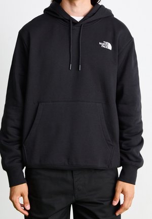 Osoba nosząca czarną bluzę z kapturem The North Face z przednią kieszenią i sznureczkami, w połączeniu z czarnymi spodniami na jednolitym tle.