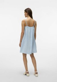 Vestido de verano de algodón a rayas azul y blanco con tirantes delgados, espalda fruncida y corte suelto, que cae por encima de las rodillas. Llevado con sandalias de tacón.