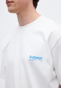 Νέος άνδρας που φοράει λευκό T-shirt με μικρό μπλε λογότυπο «lindbergh» στο αριστερό στήθος, μπροστά σε απλό φόντο.