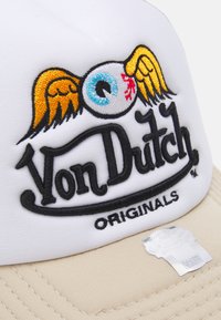 Gorra blanca con un prominente logo bordado negro y amarillo que presenta alas y un ojo. La visera es beige y tiene una tela texturizada.