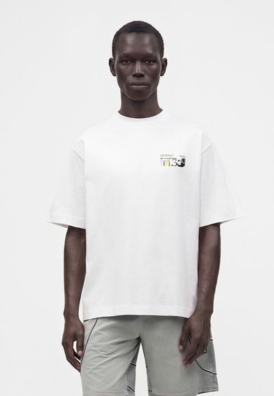 Giovane uomo con pelle scura che indossa una t-shirt bianca oversize con una piccola grafica sul petto e pantaloncini grigio chiaro su sfondo bianco.