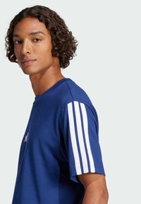 T-shirt en coton bleu avec des accents à trois rayures blanches sur les manches, un col rond et une coupe décontractée. Texture lisse et marquage minimal.