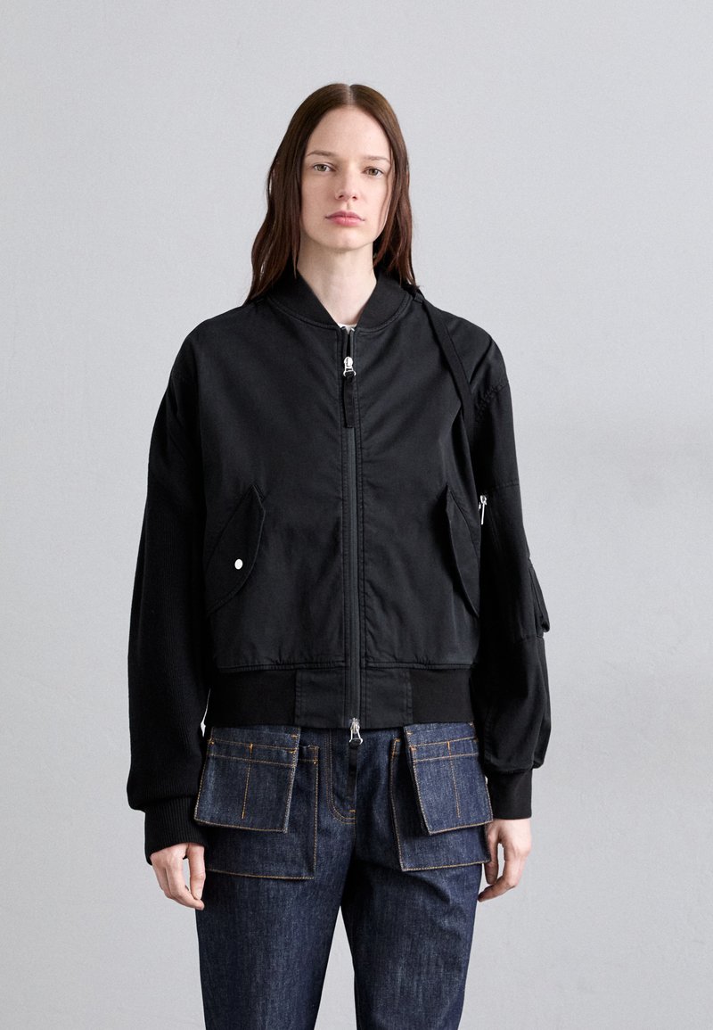 Fekete bomber dzseki elülső cipzárral, két oldalsó zsebbel és bordás mandzsettával. Kék farmernadrág felett viselve, nagy patkózsebekkel.