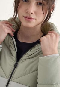Una joven con cabello oscuro y suelto aparece en primer plano, vistiendo una chaqueta acolchada de color verde claro, sosteniendo la cremallera, con un fondo neutro.