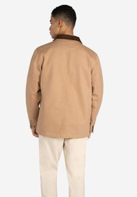 Chaqueta de lienzo marrón con cuello de pana, corte oversize y puños abotonados. Vista trasera simple, combinada con pantalones de color claro.