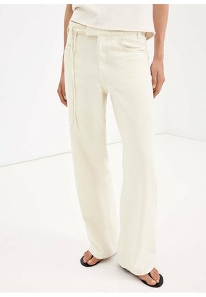 Persona che indossa pantaloni larghi color avorio con cintura annodata in vita e infradito nere, in piedi su un pavimento bianco contro una parete bianca.