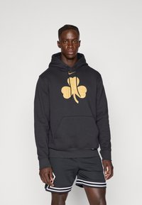 Felpa nera con un grande logo giallo a trifoglio, cappuccio con stringa e tasca frontale. Abbinata a pantaloni corti in rete nera traspirante con strisce bianche.