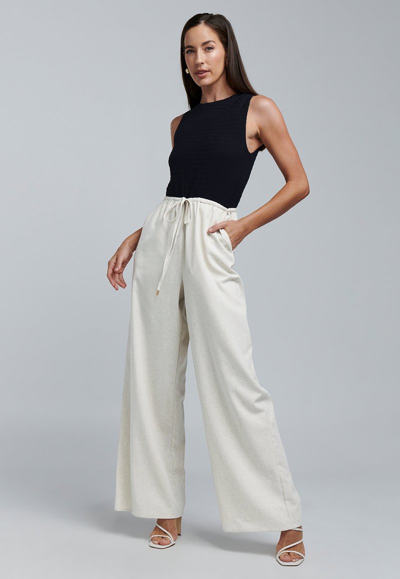 Melns bezroku tops ar teksturētu audumu un augstu apkakli, kombinēts ar krēmkrāsas platām biksēm ar elastīgu jostiņu un kabatām.