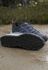 Deux chaussures de trail grises et noires avec des semelles texturées sur un chemin de gravier avec de l’herbe en arrière-plan.