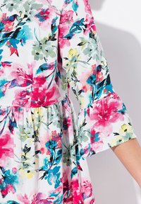 Blumenkleid mit lebendigen rosa, blauen und gelben Mustern auf weißem Hintergrund, mit Rüschärmeln und gerafftem Taillenbund.