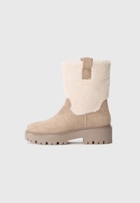 Beige suède laarzen van gemiddelde hoogte met dikke rubberen zool, voering van schapenwol aan de bovenrand en een trekband, op een neutrale grijze achtergrond.