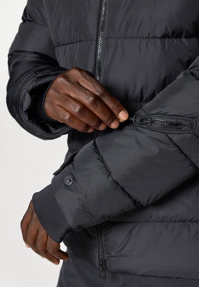 Giubbotto piumino nero con tasca a zip sul braccio, polsini a costine e finitura testurizzata. Sono visibili dettagli in zip e bottoni.
