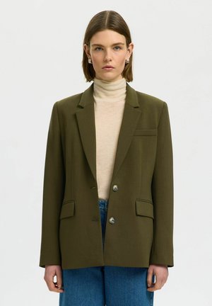 Donna con capelli castani lisci indossa un blazer verde oliva sopra un dolcevita beige e jeans blu, in piedi davanti a uno sfondo bianco semplice.