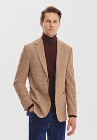 Brązowa marynarka w kratę ze złotymi guzikami, o dopasowanym kroju, noszona na ciemnym golfie; zestawiona z niebieskimi dżinsami, o gładkiej teksturze, casualowy styl.