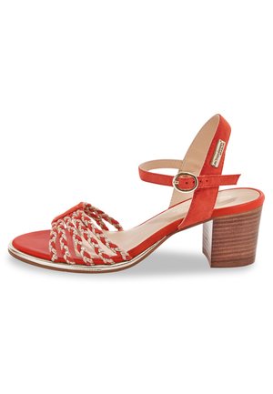 Sandale à talon en daim rouge avec un design tressé, bride à la cheville et accent doré. Le talon en bois à effet grain de bois ajoute de la stabilité et un contraste visuel.