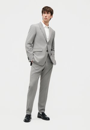 Νέος άνδρας που φοράει ανοιχτό γκρι κοστούμι, λευκό πουκάμισο και μαύρα παπούτσια, στέκεται με τα χέρια στις τσέπες κοιτώντας προς τα πλάγια.