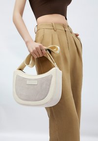 Lyscreme håndtaske med et gråt suede-panel, blød og slaskende form, og et unikt båndlignende håndtag. Båret af en person i beige bukser.