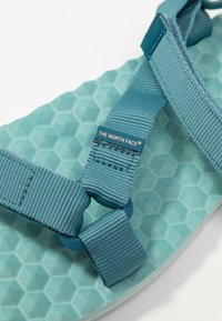 Sandal s aqua barvou, s podrážkou s texturou včelí plástve a nastavitelnými tkanými popruhy. Obsahuje štítek s logem na popruhu.