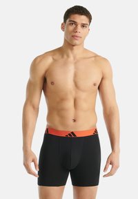 Muž bez trička stojí, má na sobě černé boxerky s oranžovým pasem, na kterém je černé logo Adidas.