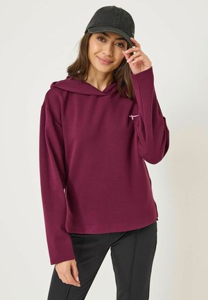 Felpa con cappuccio bordeaux realizzata in morbido tessuto, con maniche lunghe e un piccolo logo sul petto. Indossata con pantaloni neri e un berretto.