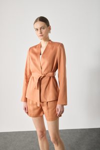 Light orange blazer with a tie waist and notched collar, paired with matching shorts. Smooth texture, structured design, and minimal detailing.
