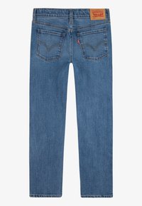 Jean en denim bleu à jambe droite, vu de dos avec deux poches arrière, un patch en cuir marron de la marque et une étiquette rouge de la marque sur la poche droite.