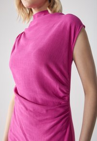 Robe fuchsia avec un col montant, des manches courtes et des fronces sur les côtés. Le tissu a une texture subtile, offrant un aspect à la fois structuré et doux.