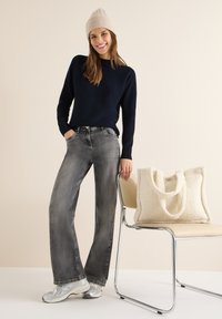 Maglione blu navy, jeans grigi a gamba larga e sneakers metallizzate. Borsa tote crema con manici morbidi appoggiata su una sedia cromata.