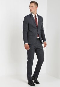 Donkergrijs, gestructureerd pak met een slim fit, gecombineerd met een wit dresshirt en een rode stropdas. Zwarte leren schoenen maken de look compleet.