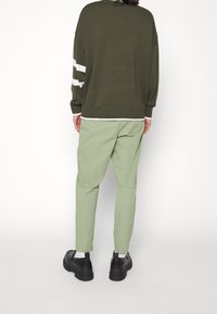 Osoba stojící zády, oblečená v olivově zeleném svetru s bílými proužky a světle zelených kalhotách, doplněná černými botami a bílými ponožkami.