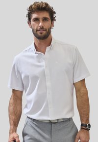 Kurzärmliges, weißes Hemd mit Button-Down-Kragen, strukturiertem Stoff und subtiler glänzender Oberfläche. Kombiniert mit hellgrauen Hosen und einer Uhr.