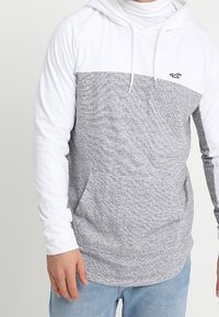 Grå och vit huvtröja med en texturerad grå kropp, vita ärmar och en framficka. Har en liten logotyp på bröstet.