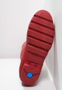 Semelle de botte en caoutchouc rouge avec des motifs en relief, comportant un accent circulaire bleu et le logo "HUNTER" en relief. Talon plat, sans lacets ni éléments apparents.