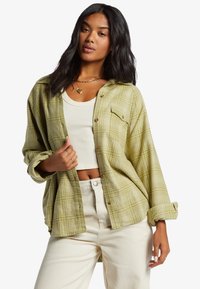 Grønternet skjorte lavet af let materiale, med knaplukning fortil, to brystlommer og opsmøgede ærmer, båret over en hvid top.