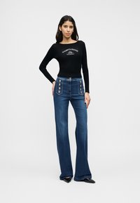 Μαύρο ριμπ μπλουζάκι με μακριά μανίκια και λευκή γραπτή λογότυπο, συνδυασμένο με σκουρόχρωμο μπλε τζιν με καμπάνα και διακοσμητικά κουμπιά στις μπροστινές τσέπες.