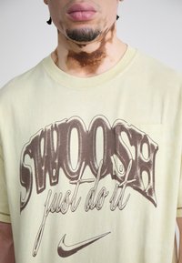 Žuta pamučna majica s velikim smešnim smešnim tekstom "SWOOSH just do it" u smešnom smešnom tonu i malim džepom. Mekana tekstura i opušteni kroj.