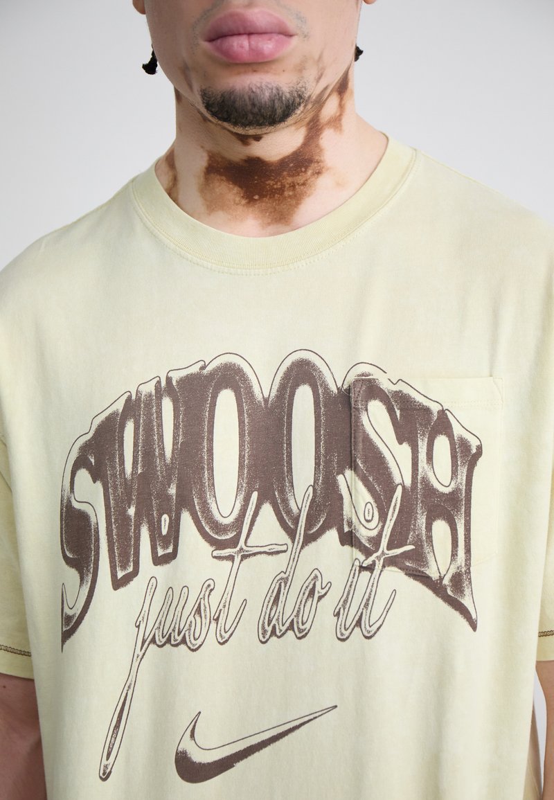 T-shirt en coton jaune avec un grand texte graphique marron "SWOOSH just do it" et une petite poche. Texture douce et coupe décontractée.