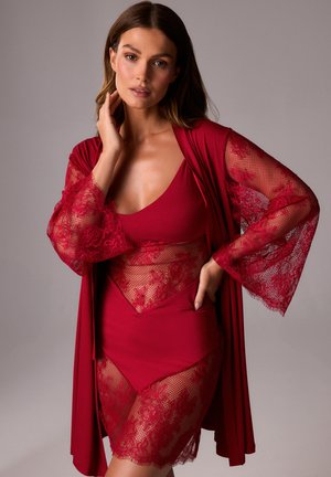 Kobieta w czerwonej koronkowej bieliźnie i pasującym szlafroku z przezroczystymi kwiatowymi rękawami, pozując z jedną ręką dotykającą szyi.