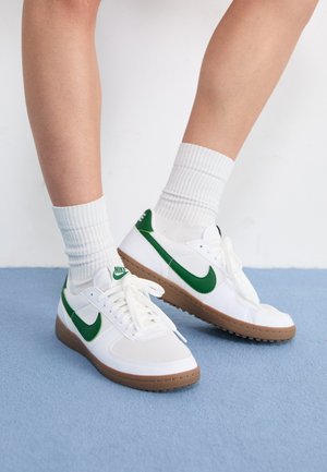 Valkoiset tennarit vihreillä yksityiskohdilla ja brändäyksellä. Kumipohja ruskealla tekstuurilla. Nauhoilla varustettu malli, yhdistetty valkoisiin ribattuihin sukkiin.