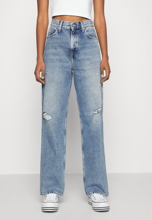 Frau trägt hellblaue, hoch taillierte, ausgewaschene Jeans mit weitem Bein, weiße Plateausneaker und ein weißes, kurz geschnittenes Oberteil.