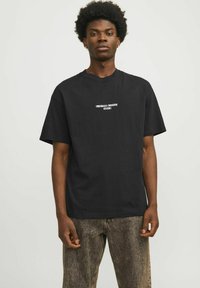 Zwarte katoenen t-shirt met korte mouwen, ronde halslijn en witte tekst "ORIGINALS CREATIVE STUDIO" prominent weergegeven op de voorkant.