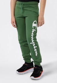 Groene joggingsbroek met wit "Champion" logo, trekkoord in de taille en taps toelopende cuffs. G gedragen met zwarte sneakers met neonaccenten.