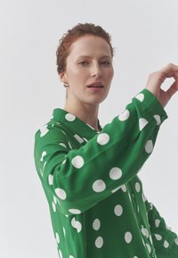 Žalia marškiniai su didelėmis baltomis taškelėmis, ilgomis rankovėmis ir apykakle. Lygi tekstūra, rodomi su modeliu, vieną ranką pakelta gestui.
