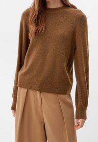 Femme portant un pull en tricot marron avec des poignets et un ourlet côtelés, associé à un pantalon plissé marron clair sur fond blanc.