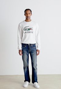 Witte sweatshirt met groen krokodillogo, gecombineerd met blauwe denim jeans en witte sneakers, staande op een neutrale achtergrond.