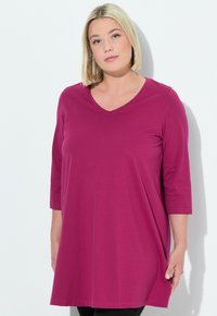 Magenta Tunika mit V-Ausschnitt und dreiviertel Ärmeln, aus weichem Stoff gefertigt. Die Länge reicht bis zur Mitte des Oberschenkels und hat einen lässigen Schnitt.