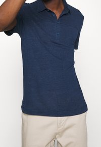 Námořnická modrá polo tričko s krátkými rukávy, límečkem a dvoubottonou légou. Obsahuje náprsní kapsu a texturovanou látku.