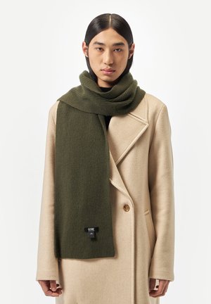Person with long black hair wears a beige wool coat and an olive green ribbed scarf with a black GOBI label on a white background.