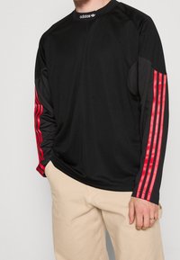 Black long-sleeve athletic shirt with red three-stripe branding on sleeves, made from smooth fabric, featuring a crew neckline and ribbed cuffs.