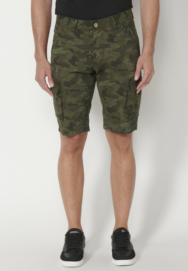 Mies, jolla on päällään vihreät camouflaged cargo-shortsit ja mustat lenkkarit, seisoo vaaleaa taustaa vasten.